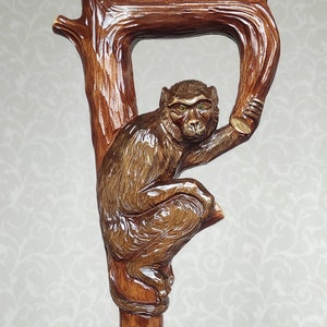 May include: A wooden walking stick with a carved monkey holding onto a branch. The monkey has brown fur and yellow eyes. The stick is brown and has a gold band around it.