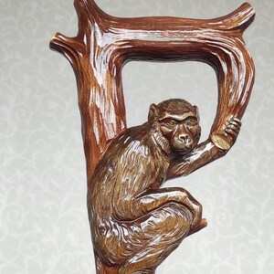 May include: A wooden walking stick with a carved monkey holding onto a branch. The stick has a brown wooden shaft with a gold band and a black band.