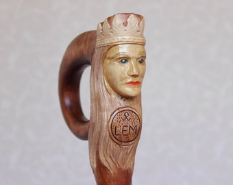 Puede incluir: Bastón de madera tallada con una cara coronada. Los ojos son azules, los labios rojos. El mango es marrón oscuro, contrastando con la madera clara de la cara. Un emblema circular con las letras "LEM" está tallado debajo.
