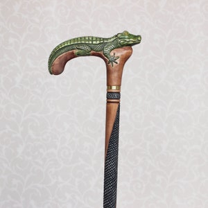 May include: A wooden walking stick with a carved alligator head handle. The handle is green and brown, and the stick is black and brown.
