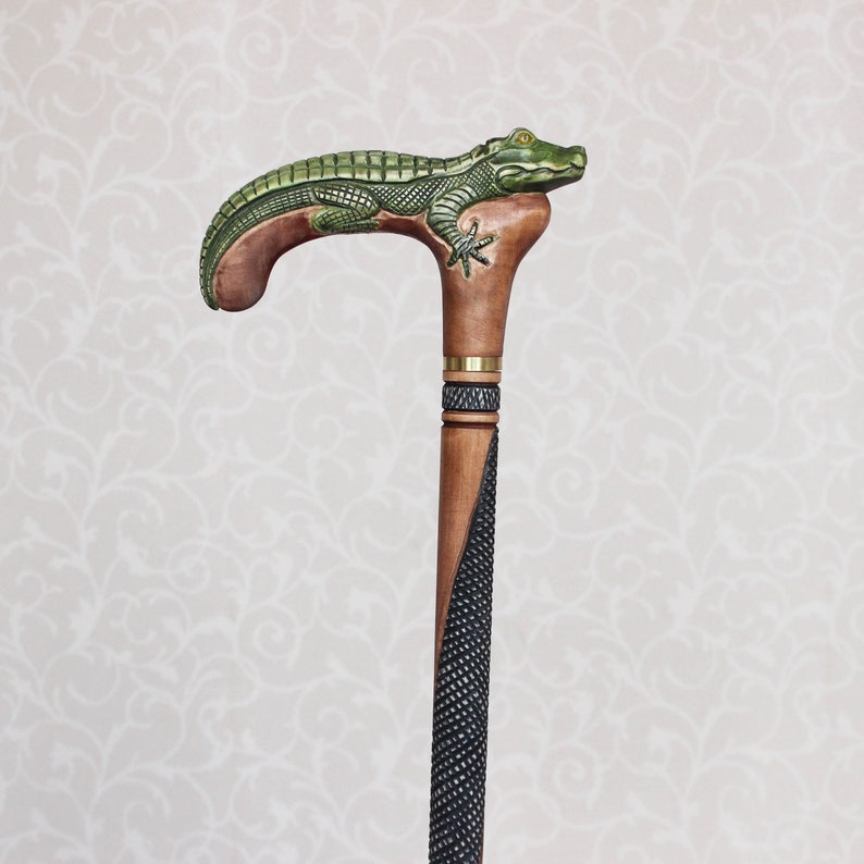 May include: A wooden walking stick with a carved alligator head handle. The handle is green and brown, and the stick is black and brown.