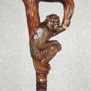 May include: A wooden walking stick with a carved monkey figure holding onto a branch. The stick has a brown finish and gold accents.