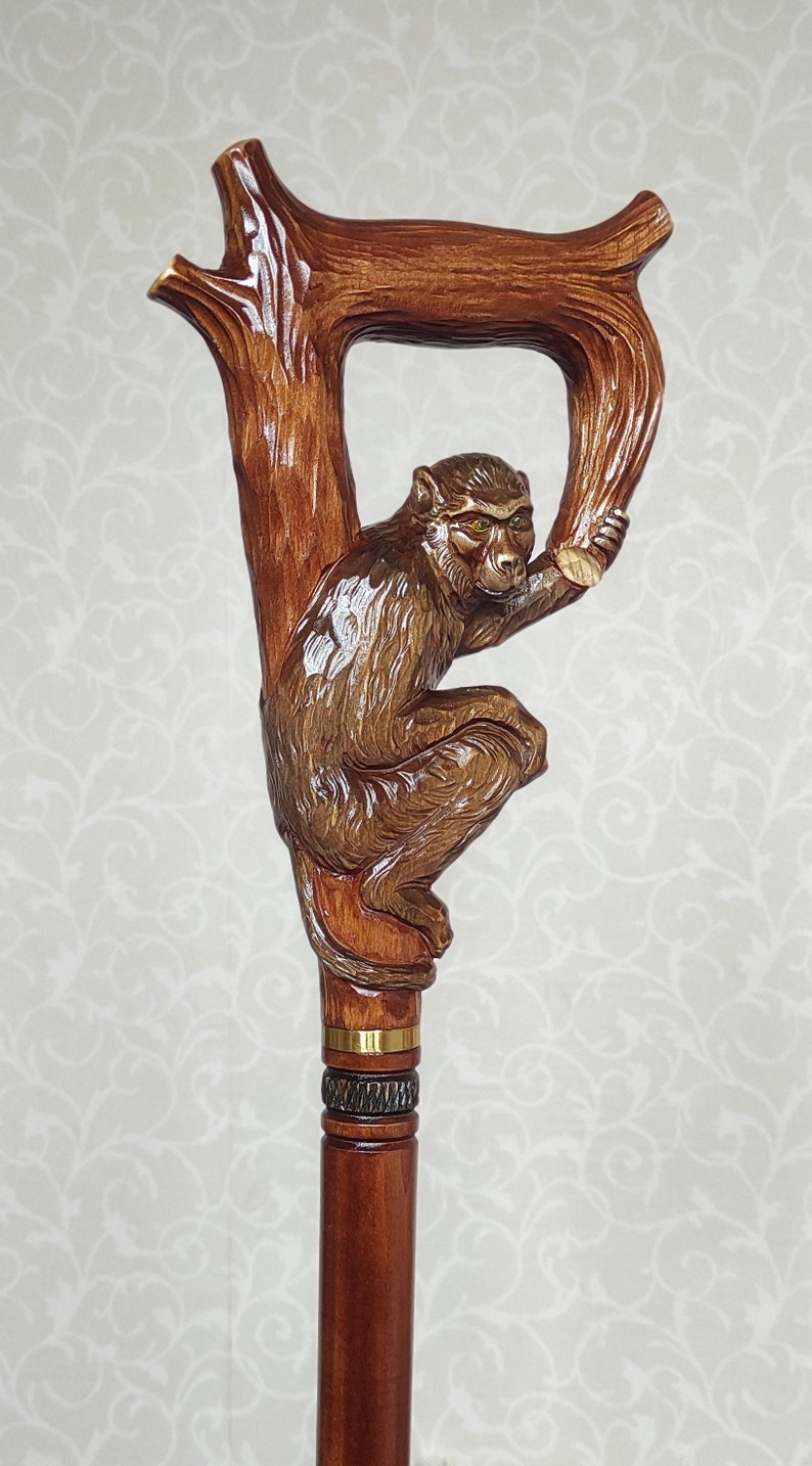 May include: A wooden walking stick with a carved monkey figure holding onto a branch. The stick has a brown finish and gold accents.