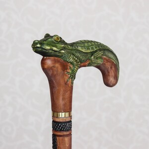 May include: A wooden walking stick with a carved alligator head handle. The alligator is green and brown with yellow eyes. The stick is brown with a gold band and a black and white patterned section.