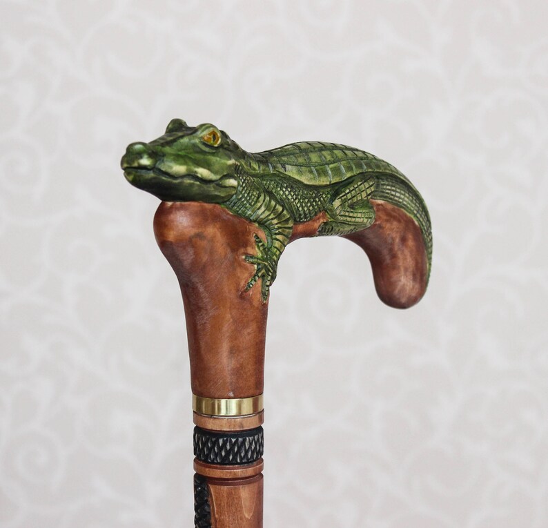 May include: A wooden walking stick with a carved alligator head handle. The alligator is green and brown with yellow eyes. The stick is brown with a gold band and a black and white patterned section.