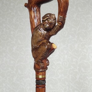 May include: A wooden walking stick with a carved monkey holding onto a branch. The stick is brown and has a gold band around it.