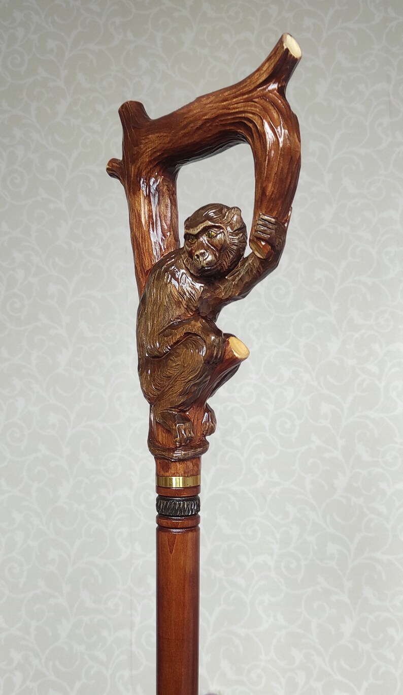 May include: A wooden walking stick with a carved monkey holding onto a branch. The stick is brown and has a gold band around it.