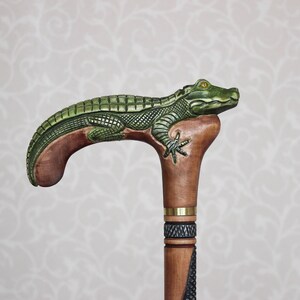 May include: A wooden walking stick with a carved alligator head and body. The alligator is green and brown and has a yellow eye. The stick is brown and has a brass band around it.