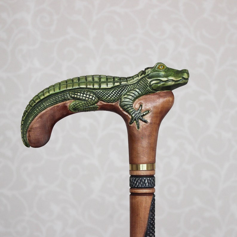 May include: A wooden walking stick with a carved alligator head and body. The alligator is green and brown and has a yellow eye. The stick is brown and has a brass band around it.