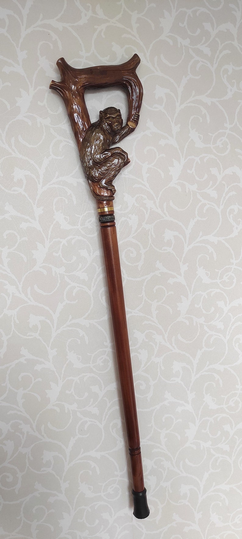 May include: A wooden walking stick with a carved monkey figure at the top. The monkey is brown and sitting on a branch. The stick is brown and has a black rubber tip.