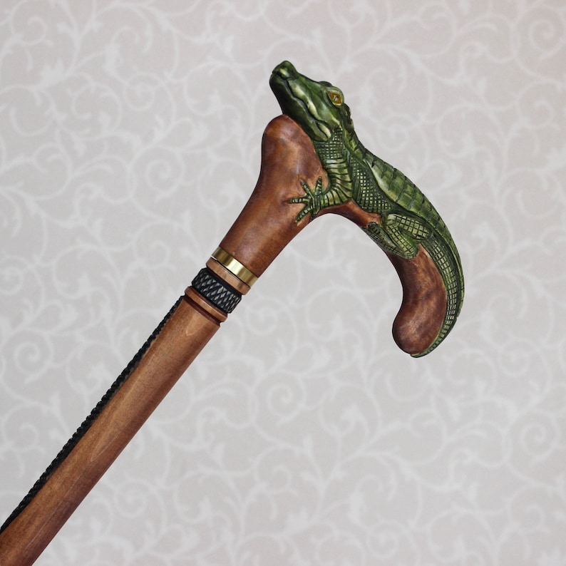May include: A wooden walking stick with a carved alligator head handle. The alligator is green and brown with a textured surface. The stick is brown with a black and gold band.