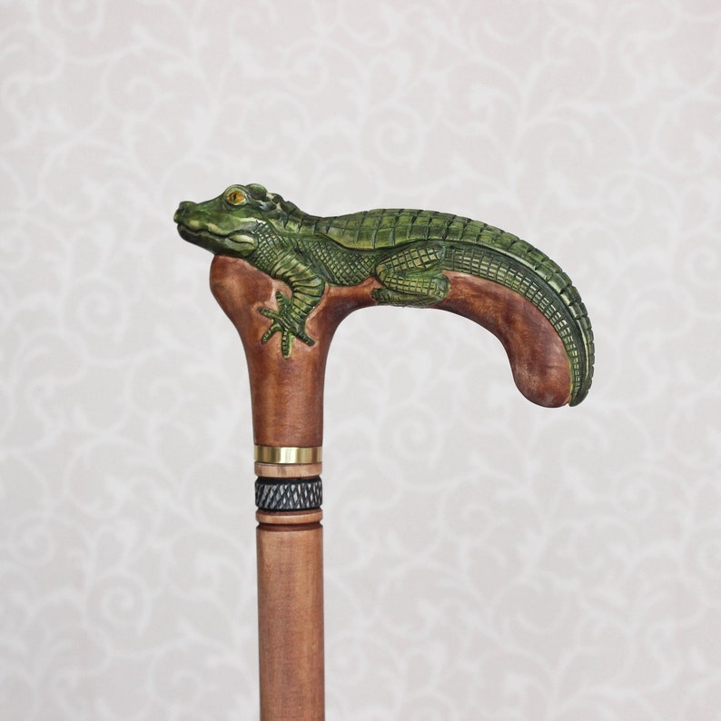 May include: A wooden walking stick with a carved alligator head handle. The alligator is green and brown with a yellow eye. The stick is brown and has a black and gold band around it.