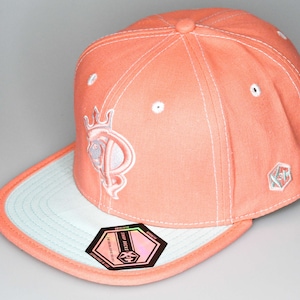 May include: A coral coloured baseball cap with a white brim and a white embroidered logo on the front. The logo features a crown and the letter "P". The cap has a white "New Era" tag on the side.