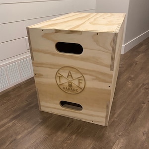 Puede incluir: Caja pliométrica de madera clara con asas recortadas. La caja presenta un logotipo circular con el texto "TAF GARAGE EST 2020". Diseñada para ejercicios y rutinas de fitness.