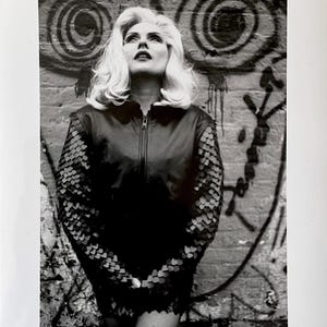 May include: Black and white photograph of a woman with blonde hair looking upwards. She wears a black jacket with scale-like detailing on the sleeves, standing in front of a graffiti-covered brick wall.