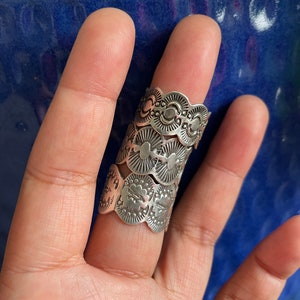 May include: A silver finger ring with an elaborate design. The ring has multiple layers with scalloped edges and embossed patterns, including floral and geometric shapes. The ring is worn on a finger.