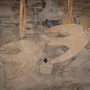May include: Two white, bird-shaped ornaments with outstretched wings hang from the top of the frame. Each bird has a small bell attached. The birds are made of a textured, open-weave material, and are suspended by a natural-colored string.