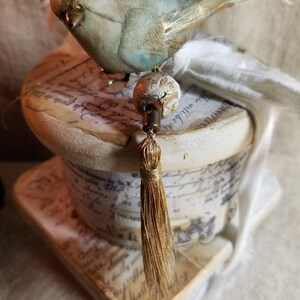 May include: A vintage-style spool with a white background and gold accents. The spool is decorated with a small blue bird and a gold tassel. The spool is sitting on a white surface.