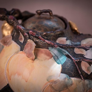 May include: A ceramic teapot with a brown, black, and gold design. The teapot is decorated with painted peach shapes and a branch with leaves.