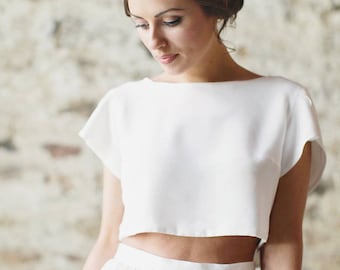 May include: A white crop top with short sleeves and a white skirt. The top has a simple, rounded neckline. The skirt is gathered at the waist and falls to the floor.