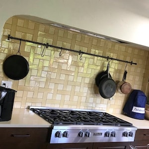 May include: A black metal pot rack with hooks hanging above a stainless steel stovetop with five burners. The stovetop is in a kitchen with beige tile backsplash.