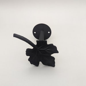 May include: Black metal leaf-shaped wall hook with a round base. The hook is attached to a wall.