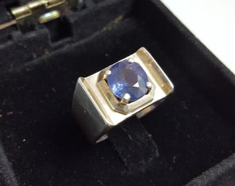 May include: A silver ring with a large, dark blue gemstone set in a square bezel. The ring is in a black velvet box.