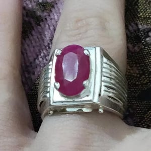 Może przedstawiać: Srebrny pierścionek z dużym, owalnym rubinem osadzonym w oprawie typu bezel. Pierścionek ma fakturowany obrączkę z szeregiem pionowych linii.