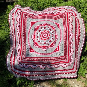 Puede incluir: Una manta de ganchillo con un gran diseño circular intrincado en el centro. La manta está hecha con una combinación de hilo rojo, rosa y gris.