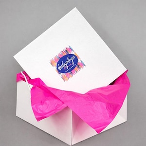 May include: A white gift box with a partially opened lid, revealing bright pink tissue paper inside. The lid features a colorful logo with the text "derbyologie" in a blue oval. The box is on a gray background.