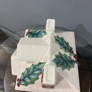 K&ouml;nnte beinhalten: Ein wei&szlig;er Keramikbeh&auml;lter in Hausform mit strukturiertem Dach und Schornstein. Das Dach ist mit gr&uuml;nen Stechpalmenbl&auml;ttern und roten Beeren verziert. Die Basis hat ein rosa Blumenmuster. Eine festliche Weihnachtsdekoration.