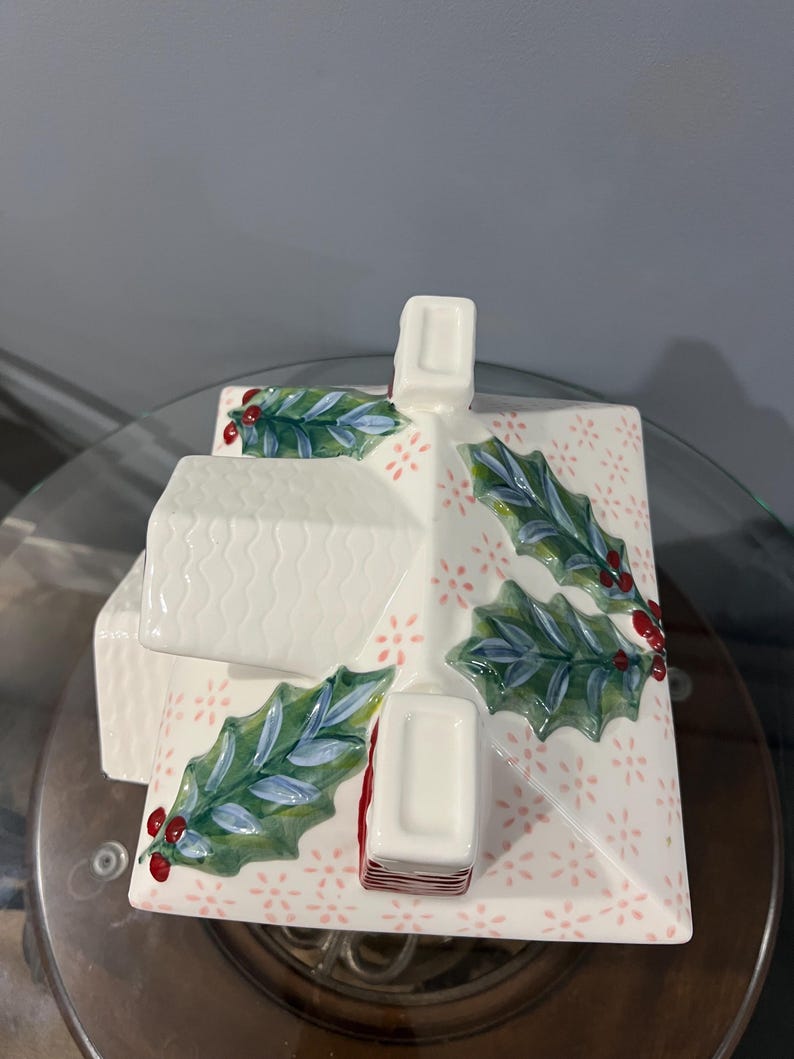 K&ouml;nnte beinhalten: Ein wei&szlig;er Keramikbeh&auml;lter in Hausform mit strukturiertem Dach und Schornstein. Das Dach ist mit gr&uuml;nen Stechpalmenbl&auml;ttern und roten Beeren verziert. Die Basis hat ein rosa Blumenmuster. Eine festliche Weihnachtsdekoration.