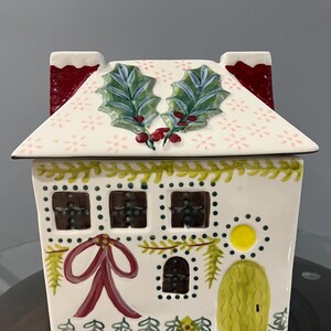 K&ouml;nnte beinhalten: Ein keramikhausf&ouml;rmiger Beh&auml;lter mit wei&szlig;em Sockel und Dach. Das Dach hat ein Blumenmuster und Stechpalmenbl&auml;tter. Das Haus hat Fenster, eine rote Schleife und eine gelbe T&uuml;r, mit einer Girlande aus gr&uuml;nen Bl&auml;ttern und Beeren.