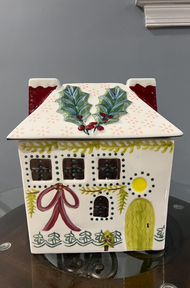 K&ouml;nnte beinhalten: Ein keramikhausf&ouml;rmiger Beh&auml;lter mit wei&szlig;em Sockel und Dach. Das Dach hat ein Blumenmuster und Stechpalmenbl&auml;tter. Das Haus hat Fenster, eine rote Schleife und eine gelbe T&uuml;r, mit einer Girlande aus gr&uuml;nen Bl&auml;ttern und Beeren.