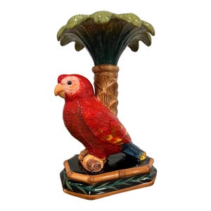 May include: A ceramic parrot figurine perched on a palm tree base. The parrot is primarily red with yellow and blue accents. The palm tree has a green frond top and a brown trunk. The base is black with bamboo detailing.