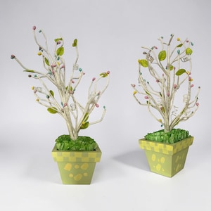 May include: Two decorative artificial trees in square green pots. The trees have white branches with colorful bead-like accents and small green leaves. The pots are light green with yellow checkered patterns and decorative spots.