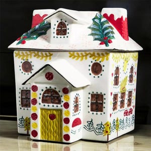 K&ouml;nnte beinhalten: Ein wei&szlig;er Keramik-Keksdose in Form eines Lebkuchenhauses. Das Haus hat einen roten Schornstein, braune Fenster und eine gelbe T&uuml;r. Das Dach ist mit gr&uuml;nem Stechpalmen und roten Beeren verziert. Die Dose ist mit farbenfrohen Details verziert.
