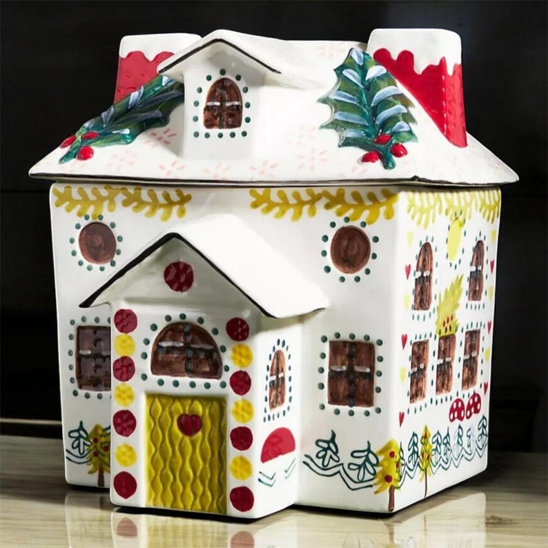 K&ouml;nnte beinhalten: Ein wei&szlig;er Keramik-Keksdose in Form eines Lebkuchenhauses. Das Haus hat einen roten Schornstein, braune Fenster und eine gelbe T&uuml;r. Das Dach ist mit gr&uuml;nem Stechpalmen und roten Beeren verziert. Die Dose ist mit farbenfrohen Details verziert.