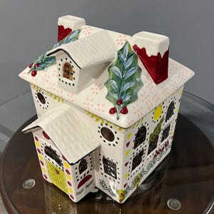 K&ouml;nnte beinhalten: Ein keramischer Lebkuchenhaus-Beh&auml;lter mit wei&szlig;em Dach und W&auml;nden, die mit roten, gelben und gr&uuml;nen Akzenten verziert sind. Das Dach hat Stechpalmen- und Beerenverzierungen, und die Schornsteine sind rot mit wei&szlig;em Rand. Das Haus hat Fenster und eine gelbe T&uuml;r.