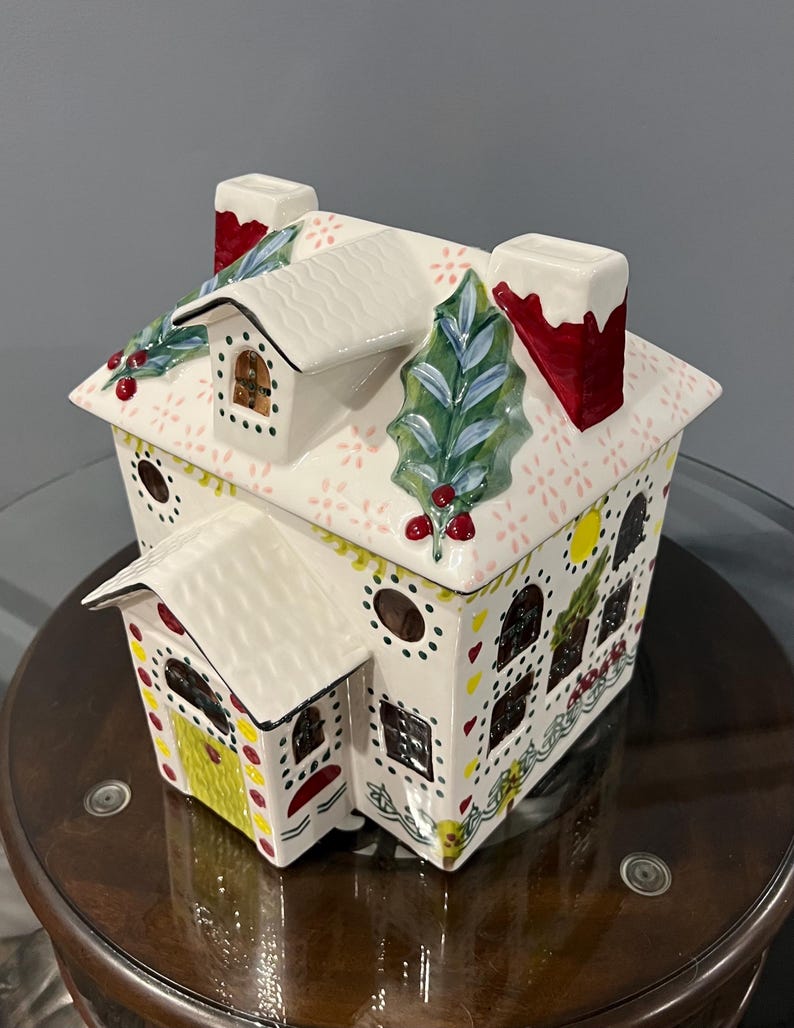 K&ouml;nnte beinhalten: Ein keramischer Lebkuchenhaus-Beh&auml;lter mit wei&szlig;em Dach und W&auml;nden, die mit roten, gelben und gr&uuml;nen Akzenten verziert sind. Das Dach hat Stechpalmen- und Beerenverzierungen, und die Schornsteine sind rot mit wei&szlig;em Rand. Das Haus hat Fenster und eine gelbe T&uuml;r.