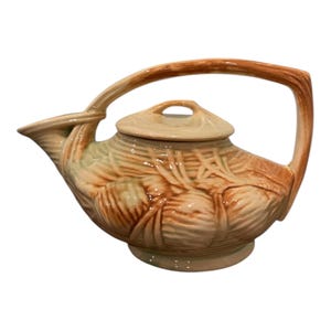 May include: A ceramic teapot with a light brown handle and lid. The body of the teapot is a light green color with a textured, carved design. The teapot has a spout and a curved handle.