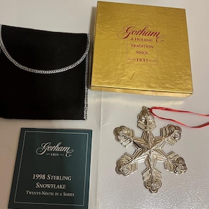 May include: A silver snowflake ornament with a red ribbon, a black pouch, a gold box with the text "Gorham A Holiday Tradition Since 1831", and a card that reads "1998 Sterling Snowflake Twenty-Ninth in a Series."