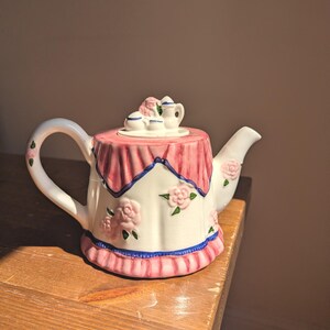 May include: A ceramic teapot shaped like a table with a pink tablecloth, featuring raised pink rose floral accents, a blue trim along the base and top, and a lid topped with a miniature tea set including a tiny teapot and two cups.