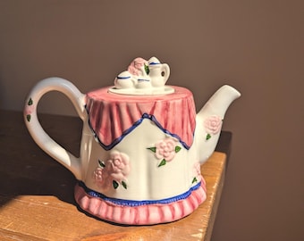 May include: A ceramic teapot shaped like a table with a pink tablecloth, featuring raised pink rose floral accents, a blue trim along the base and top, and a lid topped with a miniature tea set including a tiny teapot and two cups.