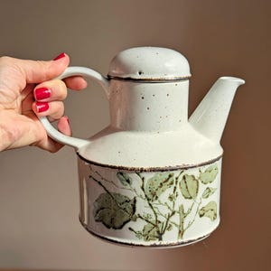 May include: A white ceramic teapot with a speckled finish and a brown rim. The body features a dark green botanical illustration of leaves and branches. It has a cylindrical base, a curved handle, a matching lid, and a pointed spout.