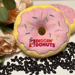 May include: A pink and yellow donut-shaped plush toy with the text "Diggin' Donuts" and sprinkles. The toy is sitting on a surface with coffee beans.