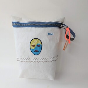 Könnte beinhalten: Weiße Tasche mit blauem Reißverschluss und blau-oranger Handschlaufe. Die Tasche hat ein gesticktes Gesichtsdesign in Gelb, Blau und Rot, mit den Initialen "R.E.H." über dem Design. Unten ist eine braune Zickzacknaht aufgenäht.