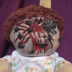 May include: A creepy doll with a red and white striped face covered in fake spiders and blood. The doll is wearing a white shirt with a colorful pattern of birds.