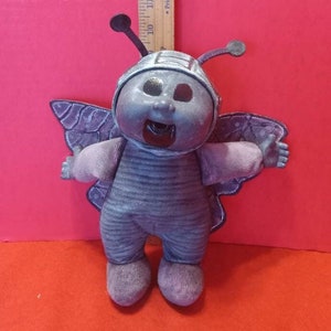 May include: A gray plush toy shaped like a butterfly with purple wings. The toy has a smiling face with large eyes and a wide open mouth.
