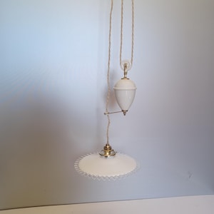 Puede incluir: Lámpara colgante blanca con pantalla festoneada y fijación de latón. La lámpara cuelga de un sistema de poleas con un peso de cerámica blanca. El cable es de color beige trenzado.